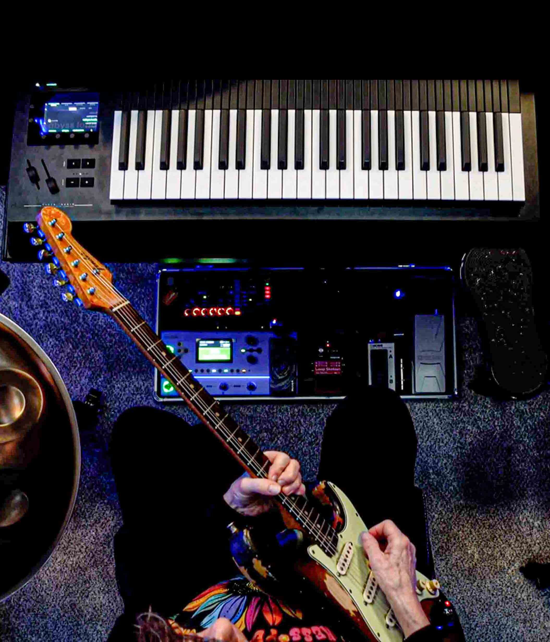 Kelly Richey playing electric guitar in a home studio with MIDI keyboard, effects pedals, and music production equipment during creative session