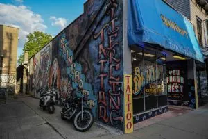 Colorful corner tattoo and piercing shop with blue awning, graffiti-covered side wall, and motorcycles parked outside in an urban alley setting.