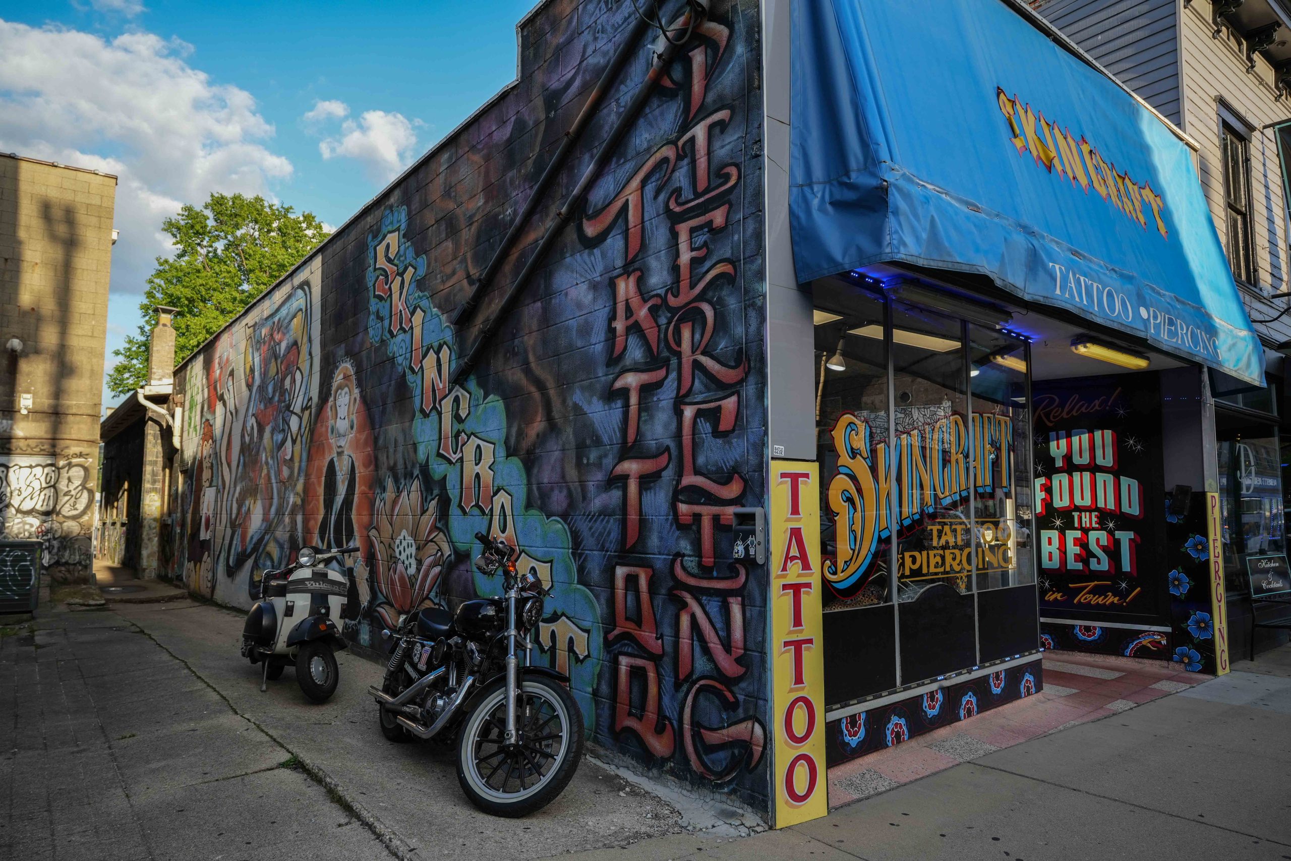 Colorful corner tattoo and piercing shop with blue awning, graffiti-covered side wall, and motorcycles parked outside in an urban alley setting.